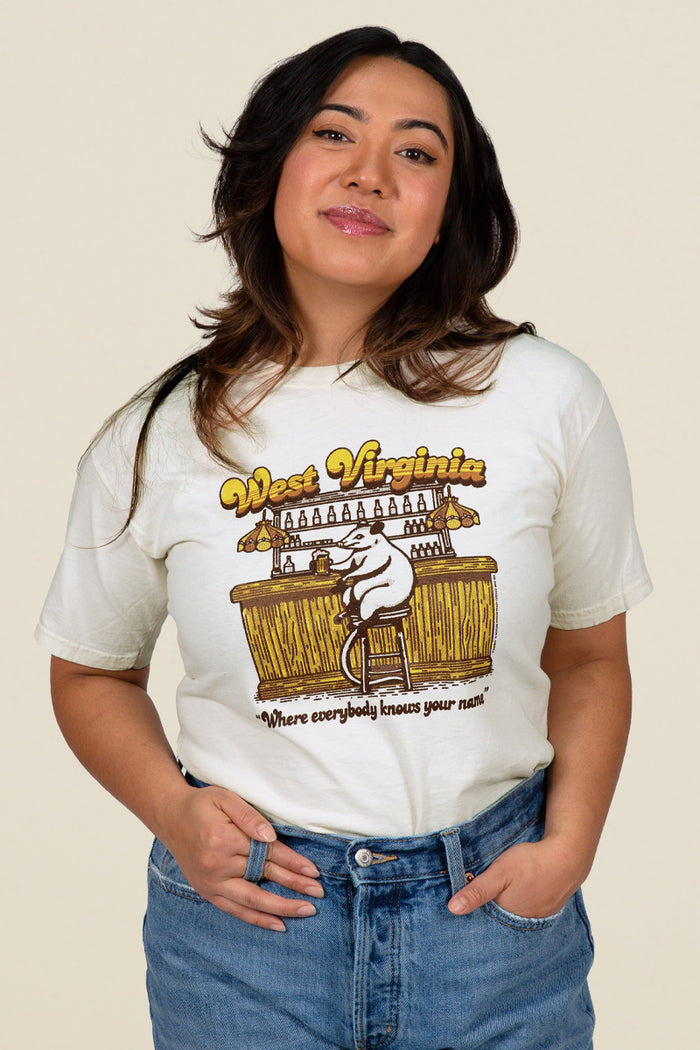 Kin Ship Goods cream tee with opossum at a bar illustration, retro ‘West Virginia’ text, and ‘Where everybody knows your name’ slogan.