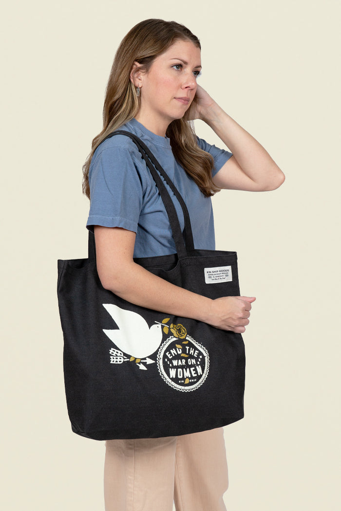 Kin Ship Goods black tote bag with white dove, gold rose, and 'End the War on Women' message in bold black and white circle.