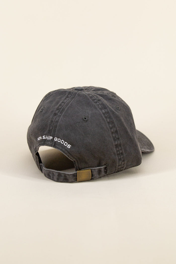 Black Kin Ship Goods baseball cap with white lantern embroidery on front, laid at an angle on neutral background.