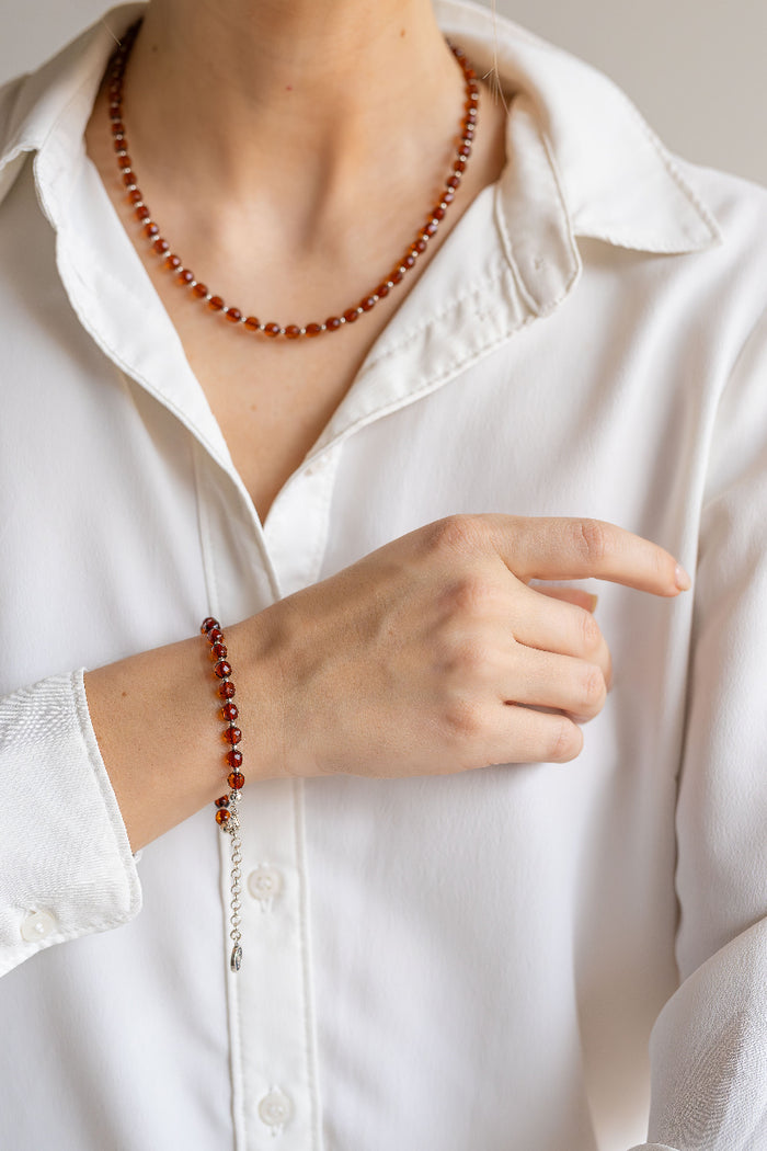 Person wearing a white shirt with a beaded amber necklace and bracelet, pointing to the jewelry.