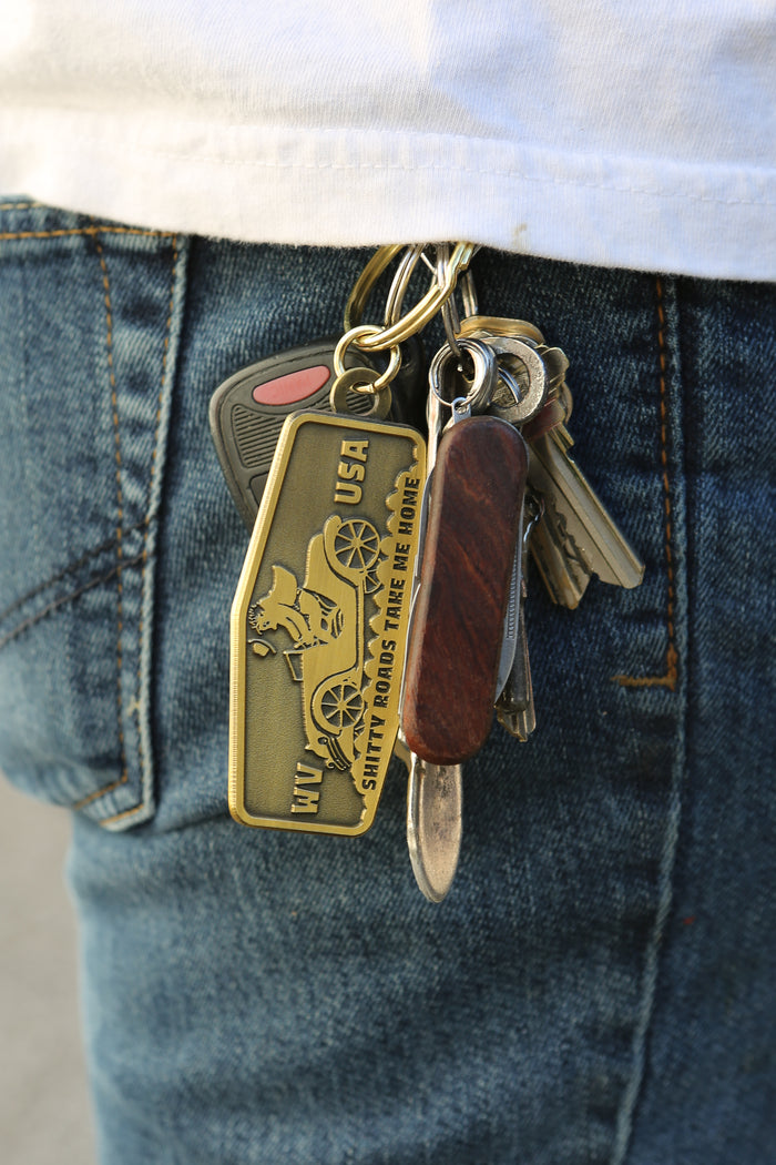 Brass coffin-shaped keychain from Kin Ship Goods with vintage car and “Shitty Roads Take Me Home” design, inspired by West Virginia road trips.