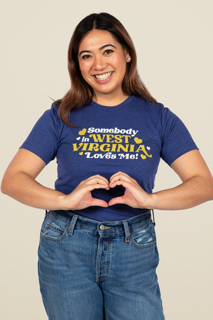Kin Ship Goods adult tee in heather blue with gold and white ‘Somebody in West Virginia Loves Me!’ retro-style graphic.